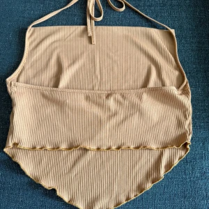 Brun ribbad halterneck linnetopp - Supersnygg brun ribbad linnetopp med halterneck och knyt i nacken. Toppen har en croppad passform med vågig nederkant och smala axelband. Perfekt för varma dagar och ger en trendig vibe till din outfit.