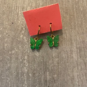 Gröna fjärilsörhängen i guldton - Superfina örhängen med gröna fjärilar och guldfärgade detaljer. Hängena har en snygg metallisk finish och fjärilarna är i en klar grön nyans. Baksidan är texturerad i guld. Perfekta för dig som vill sticka ut med något lekfullt och trendigt.