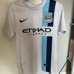 Manchester City Aguero bortatröja Nike - Manchester City bortatröja med Aguero 16 på ryggen. Vit bas med två vertikala ränder i ljusblått och marinblått, klubbmärke och Nike-logga på bröstet. Tillverkad i lätt och ventilerande Dri-FIT-material, klassisk passform och korta ärmar.