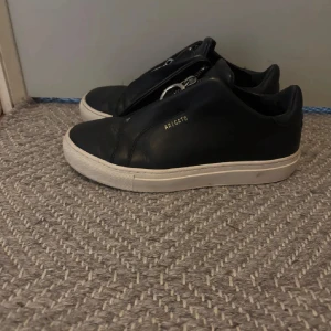 Axel Arigato skor - Stilrena svarta sneakers från Axel Arigato med vit platt sula och minimalistisk design. Skorna är i skinn och har diskret guldfärgad logga på sidan. Perfekta för dig som gillar en clean och modern look.