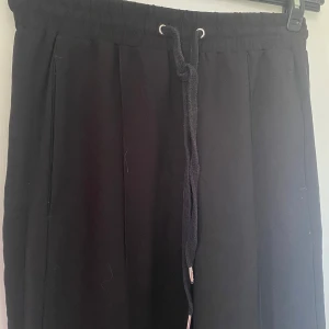 Svarta vida kostymbyxor med snörning - Svarta kostymbyxor med breda ben och hög midja. De har resår i midjan och snörning framtill med metalländar. Materialet känns mjukt och lite flowy, troligen polyester. Byxorna har fickor  på sidorna. Dragkedja på byxbenen Perfekta för en chill och stilren look.