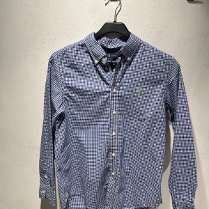 Blårutig skjorta från Ralph Lauren - Snygg blårutig skjorta från Ralph Lauren med klassisk button-down krage och vita knappar. Skjortan har lång ärm och en liten grön broderad logga på bröstet. Storlek L (14-16 år)