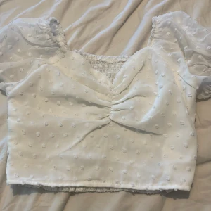 Vit croppad blus med puffärm - Superfin vit blus med croppad passform och puffiga korta ärmar. Tyget är lätt och har små broderade prickar över hela toppen. Söt rynkdetalj framtill och smockad rygg för extra komfort. Perfekt för en somrig look!