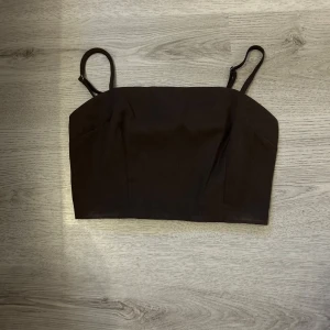 Brun croppad linnetopp med smala band - Snygg brun croppad linnetopp  med smala justerbara axelband. Toppen har en stilren, rak skärning framtill och smockad baksida för extra komfort. Perfekt att matcha med jeans eller kjol för en trendig look.