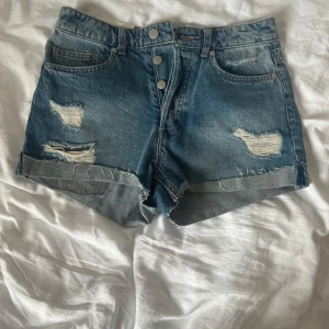 Högmidjade slitna blå jeansshorts H&M - Snygga högmidjade jeansshorts från H&M i klassisk blå denim. Shortsen har slitna detaljer, råa kanter och uppvikta benslut. Stängs med tre silverfärgade knappar framtill och har femficksmodell. Perfekta för sommardagar och festivaler