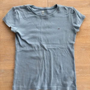 Blå t-shirt från Tommy Hilfiger - Snygg blå t-shirt från Tommy Hilfiger med klassisk rund hals och liten broderad logga på bröstet. Modellen är figurnära och har korta ärmar. Tillverkad i mjuk bomull som känns skön mot huden. Perfekt för en clean och stilren look barnstorlek.