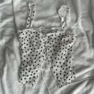 Vit prickig knyt topp från Zara - Säljer en söt vit  topp från Zara med svarta prickar och snörning framtill. Toppen har smala axelband och fransiga kanter för en trendig look. Perfekt att styla till jeans eller kjol.