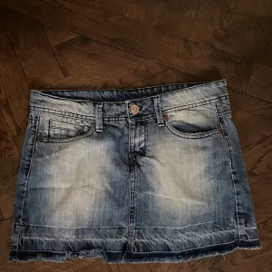 Blå jeanskjol med fransig kant - Snygg kort jeanskjol i blått med slitna detaljer och fransig nederkant. Klassisk femficksmodell med bälteshällor och knapp framtill. Perfekt för dig som gillar en avslappnad och trendig stil.