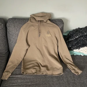 Beige/Grön  hoodie från Morris med huva - Snygg beige hoodie från Morris med broderad logga på bröstet och justerbar huva med snörning. Tröjan har långa ärmar och ribbade muddar. Perfekt för en chill och stilren look.