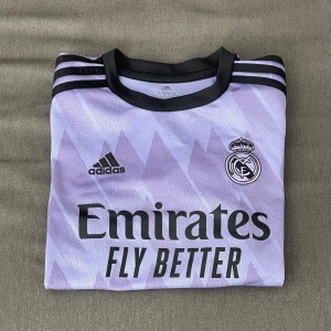 Real Madrid Adidas fotbollströja lila - Real Madrid t- shirt i snygg lila färg, änvänd fåtal gånger och är i väldigt bra skick, köpt för 999kr och är 100% äkta. Den är limiterad och är ganska svår att få tag i. Om ni har frågor är de bara att fråga på! (Svarar snabbt) priset kan diskuteras!