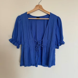 Blå blus med knyt från Gina Tricot - Blå blus i storlek XS. Den har korta puffärmar med volangkant, djup v-ringning och knyt framtill i midjan. Materialet är strukturerat och ger en luftig känsla, perfekt för vår och sommar.