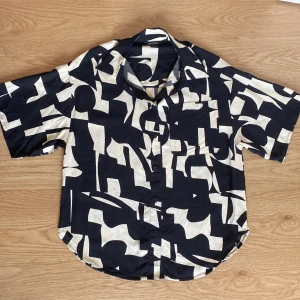 Svartvit oversize skjorta från Monki storlek XXS - Cool skjorta från Monki med svart och vit abstrakt mönster i storlek XXS men som är oversize och därmed passar både XS och S. Skjortan har korta ärmar, klassisk krage, bröstficka och knäppning framtill. Perfekt för dig som gillar statement-plagg och vill sticka ut lite extra.