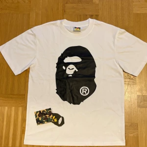 Vit BAPE t-shirt med apehuvud - Snygg vit t-shirt från BAPE med stort svart apehuvud-tryck på både fram- och baksidan. Klassisk crewneck och korta ärmar. Materialet är mjuk bomull och det finns en camo-tag med BAPE-logga vid nederkanten. Perfekt för dig som gillar streetwear.