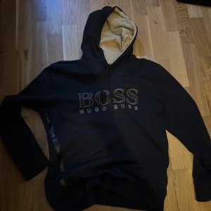 Svart hoodie från Hugo Boss - Svart hoodie från Hugo Boss med stor logga framtill och beige insida i huvan. Tröjan har långa ärmar, snörning i huvan och dekorativt band med BOSS-tryck längs sidan. Mjuk och skön bomullskänsla, perfekt för chill dagar.