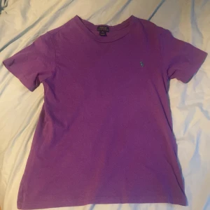 Lila t-shirt från Polo Ralph Lauren - Säljer en lila t-shirt från Polo Ralph Lauren med klassisk passform och korta ärmar. T-shirten har det ikoniska gröna broderade Polo-loggan på bröstet och är gjord i mjuk bomull. Perfekt för dig som gillar stilrena och färgglada plagg.