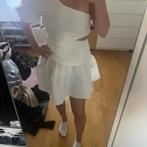 Vit one shoulder kortklänning - Superfin vit kortklänning med one shoulder och voluminös ärm. Klänningen har en markerad midja, cut out-detalj i sidan och en lätt utställd kjol. Perfekt för dig som vill sticka ut med en modern och trendig look.