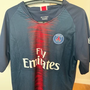 PSG Neymar Jr 10 matchtröja blå/röd - Säljer en PSG fotbollströja med Neymar Jr och nummer 10 på ryggen. Tröjan är marinblå med röda detaljer och PSG-emblem på bröstet. Fly Emirates-logga på framsidan. Tillverkad i lätt och ventilerande material, perfekt för fotboll eller supporterliv.