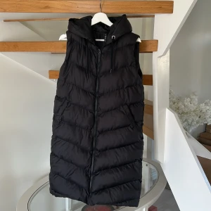 Svart lång dunväst med huva - Säljer en svart lång dunväst med huva och dragkedja framtill. Västen har snedställda fickor och quiltad design i breda, diagonala sektioner. Perfekt för lager-på-lager och chill stil under kyliga dagar.