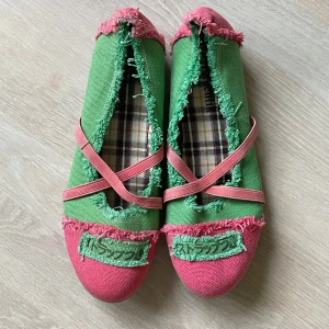 Gröna och rosa ballerinaskor med fransar - Unika ballerinaskor i grönt och rosa med fransiga kanter och coola detaljer. Skorna har korsade rosa band över foten och en rutmönstrad innersula. Tillverkade i textilmaterial med platt sula, perfekt för dig som gillar färg och statement-stil. (Ena bandet är lite missfärgat längst ner, se bild 5)