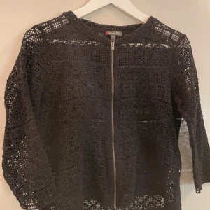 Svart stickad jacka från Street One - Svart stickad tröja från Street One med coolt genombrutet mönster och dragkedja framtill. Jackan har rund halsringning och trekvartsärm, perfekt för lager-på-lager och en chill vibe. Passar dig som gillar unika detaljer och vill sticka ut lite extra.