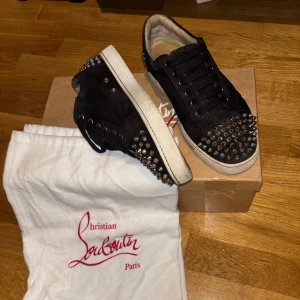 Christian Louboutin svarta sneakers med nitar - Svarta sneakers från Christian Louboutin i mocka med ikoniska röda sulor. Skorna har silverfärgade nitar på tån och hälen, svarta skosnören och vit sula. Snygg och edgy design som sticker ut, perfekt för dig som gillar statement-detaljer.