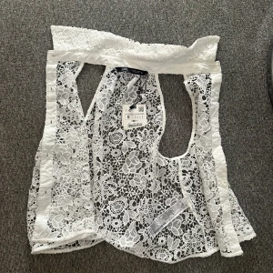 Vit spetsblus från Zara med krage - Superfin vit blus från Zara i genombruten spets med blommigt mönster. Blusen har klassisk krage, knappar bak och är ärmlös. Perfekt för dig som vill ha en romantisk och trendig look med mycket detaljer.