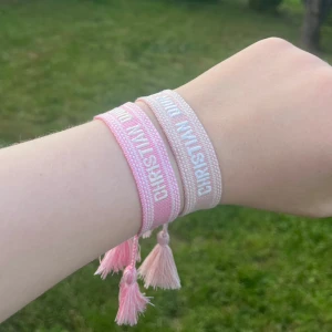 Christian Dior rosa armband med tofsar - Två snygga armband från Christian Dior i olika nyanser av rosa med vit brodyr och text. Båda har trendiga tofsar i ändarna och är perfekta för att ge din outfit en lyxig touch. Säljs tillsammans eller separat. Passar herr och dam.