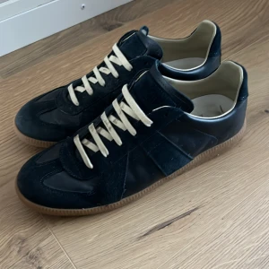  Maison Margiela  - Maison Margiela sneakers mörk bli  skinn och mocka med beige snörning och klassisk gummisula. Snygg retrostil med låg profil och rund tå. Perfekta för dig som gillar minimalistisk och tidlös design. Perfekt nu till sommaren! Priset är inte hugget i sten!