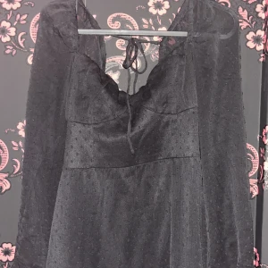 Svart kortklänning med knyt och volang - Supersöt svart kortklänning med långa ärmar och knyt i halsen. Klänningen har lätt transparent tyg med diskret prickigt mönster och volang nertill. Perfekt för dig som gillar en romantisk och trendig stil.