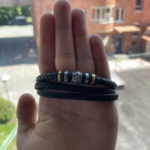 Svart flätat armband med detaljer - Stilrent svart armband i flätad design med flera varv runt handleden. Pryds av metallringar i silver och guld samt en snyggt mönstrad mittdel. Magnetlås i svart metall för enkel användning. Perfekt accessoar för en cool och modern look. Pris kan diskuteras!