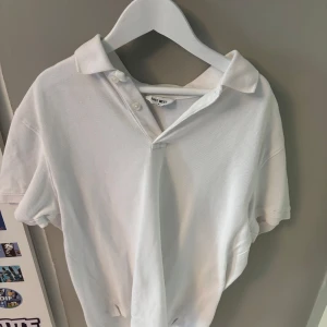Vit pikétröja från East West - Klassisk vit pikétröja från East West i storlek S. Tröjan har korta ärmar, krage och knäppning med två knappar framtill. Tillverkad i bomull för en skön och luftig känsla. Perfekt för en clean och stilren look.