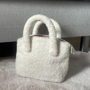 Vit fluffig handväska med axelrem - Supergullig vit handväska i mjukt teddy-material med både handtag och avtagbar axelrem. Väskan har dragkedja upptill och ett rosa innerfoder. 