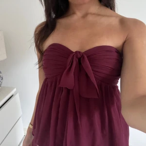 Vinröd bandeau topp med knyt - Supersöt vinröd bandeau topp med knyt framtill. Toppen är axelbandslös och har en flowy, luftig passform nedtill. Perfekt för dig som gillar en romantisk och trendig look. Materialet känns lätt och svalt, perfekt för varma dagar.