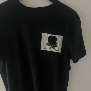 Kent & Curwen T-shirt - Svart t-shirt med från Kent & Curwen med vit patch på bröstet där en broderad mörk ros och gröna blad syns, samt texten 'K.C. 1926'. Klassisk rund hals och korta ärmar. Perfekt för dig som gillar unika detaljer och stilren design. Knappt använd 