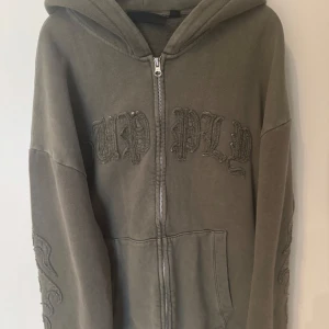 Olivgrön hoodie Supply & Demand - Olivgrön hoodie från Supply & Demand med broderad text på bröstet och detaljer på ärmarna. Tröjan har dragkedja framtill, huva och känguruficka. Oversized passform som ger en chill vibe, perfekt till streetwear-looken.