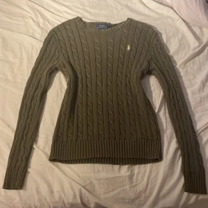 Mörkgrön Tröja Polo Ralph Lauren - Säljer en olivgrön kabelstickad tröja från Polo Ralph Lauren. Tröjan har rund halsringning, lång ärm och klassisk broderad logga på bröstet. Den är ribbad i muddar och nederkant och har en slim passform. Perfekt för lager-på-lager under hösten och vintern.