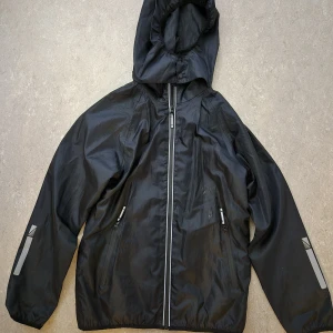 Regnjacka - Säljer en svart regnjacka från Mywear med huva och dragkedja. Jackan har långa ärmar och reflexdetaljer på ärmarna och ryggen. Finns fläck på framsidan. Perfekt för regniga dagar och utomhusaktiviteter.