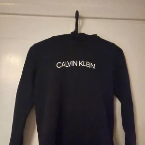 Svart hoodie från Calvin Klein Jeans - Svart hoodie från Calvin Klein Jeans med vit logga framtill. Klassisk modell med huva och långa ärmar. Perfekt för en chill och stilren look. Mjuk och skön, passar till det mesta. Snygg och enkel design som funkar året runt.