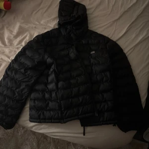 Svart dunjacka från Levi's med huva - Säljer en svart dunjacka från Levi's i storlek L. Jackan har en quiltad design, huva och dragkedja framtill. Perfekt för kalla dagar och riktigt skön att bära. Klassisk Levi's-logga på bröstet och snygg, enkel look.