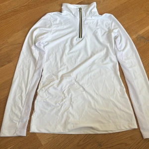 Vit långärmad ridtröja - Vit långärmad tröja i syntetmaterial med guldig half zip-dragkedja framtill och hög krage. Snyggt präglat hästmotiv på framsidan och meshpaneler längs ärmarna för extra ventilation. Perfekt för dig som gillar ridning och sportig stil.