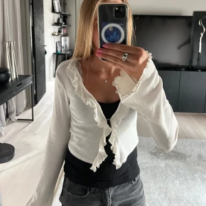 Vit omlottkofta med volangdetaljer - Supersöt vit omlottkofta med volangkant längs hela öppningen och vid ärmsluten. Koftan är ribbad och har långa ärmar med lätt utsvängd form. Står st S, men är liten i storleken så skulle säga xs. Hör av dig om du har några frågor eller funderingar!🩷