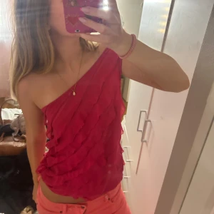 Rosa oneshoulder topp med volanger - Säljer en snygg rosa oneshoulder topp med volangdetaljer över hela framsidan.