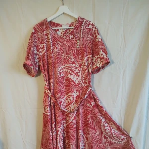 Rödvit klänning med paisleymönster - Superfin röd och vit klänning med paisleyinspirerat mönster och snygga detaljer som knappar framtill och bälte i midjan. Klänningen har korta ärmar och är tillverkad i ett mjukt, lätt material som känns skönt mot huden. Perfekt för dig som gillar färg och mönster!