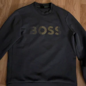 Svart sweatshirt från BOSS - Säljer en svart sweatshirt från BOSS med guldig logga framtill. Tröjan har rund halsringning och långa ärmar. Perfekt för dig som gillar stilrena och snygga plagg med en touch av lyx.