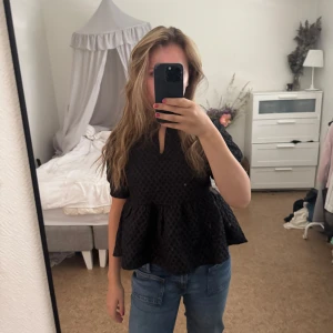 Svart blus med puffärm och struktur - Supersnygg svart blus med puffiga korta ärmar och volang nertill. Blusen har ett diskret rutigt strukturmönster och en liten v-ringning framtill. Perfekt att styla med jeans eller kjol, använt en gång. 