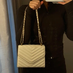 Vit axelväska med kedja - Säljer en axelväska i vitt med guldfärgad kedja och läderdetaljer. Väskan har ett klassiskt kuvertformat lock och snygga diagonala sömmar. Perfekt för dig som gillar en clean och trendig look.
