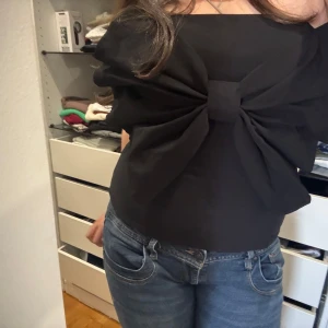 Svart tubtopp med stor rosett - Supersnygg svart tubtopp med en stor, iögonfallande rosett framtill. Toppen är axelbandslös och har en tight passform som framhäver siluetten. Perfekt för dig som vill sticka ut och skapa en cool look.