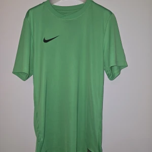 Grön Nike Dri-Fit fotbollströja S/M - NYSKICK. Snygg grön fotbollströja från Nike i Dri-Fit-material som andas. Tröjan har korta ärmar, rund hals och svart Nike-logga på bröstet. Perfekt för träning eller match, med meshpaneler på sidorna för extra ventilation.
