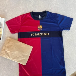 FC Barcelona fotbollströja blå/röd - Snygg FC Barcelona fotbollströja i klassiska blå och röda färger med klubbens emblem och texten 'FC BARCELONA' på bröstet. Tröjan är kortärmad och tillverkad i ett lätt och glansigt material som passar perfekt för fotbollsträning eller match. Har inte blivit använd.