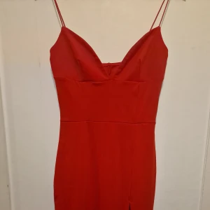 Röd klänning med smala axelband - Snygg röd klänning med smala axelband och v-ringad urringning. Modellen är figurnära och har en slits framtill för extra edge. Tvättad vid flytt och märket fastnade med bikinisnören så fick klippa bort det. Se sista bilden. Aldrig använd.
